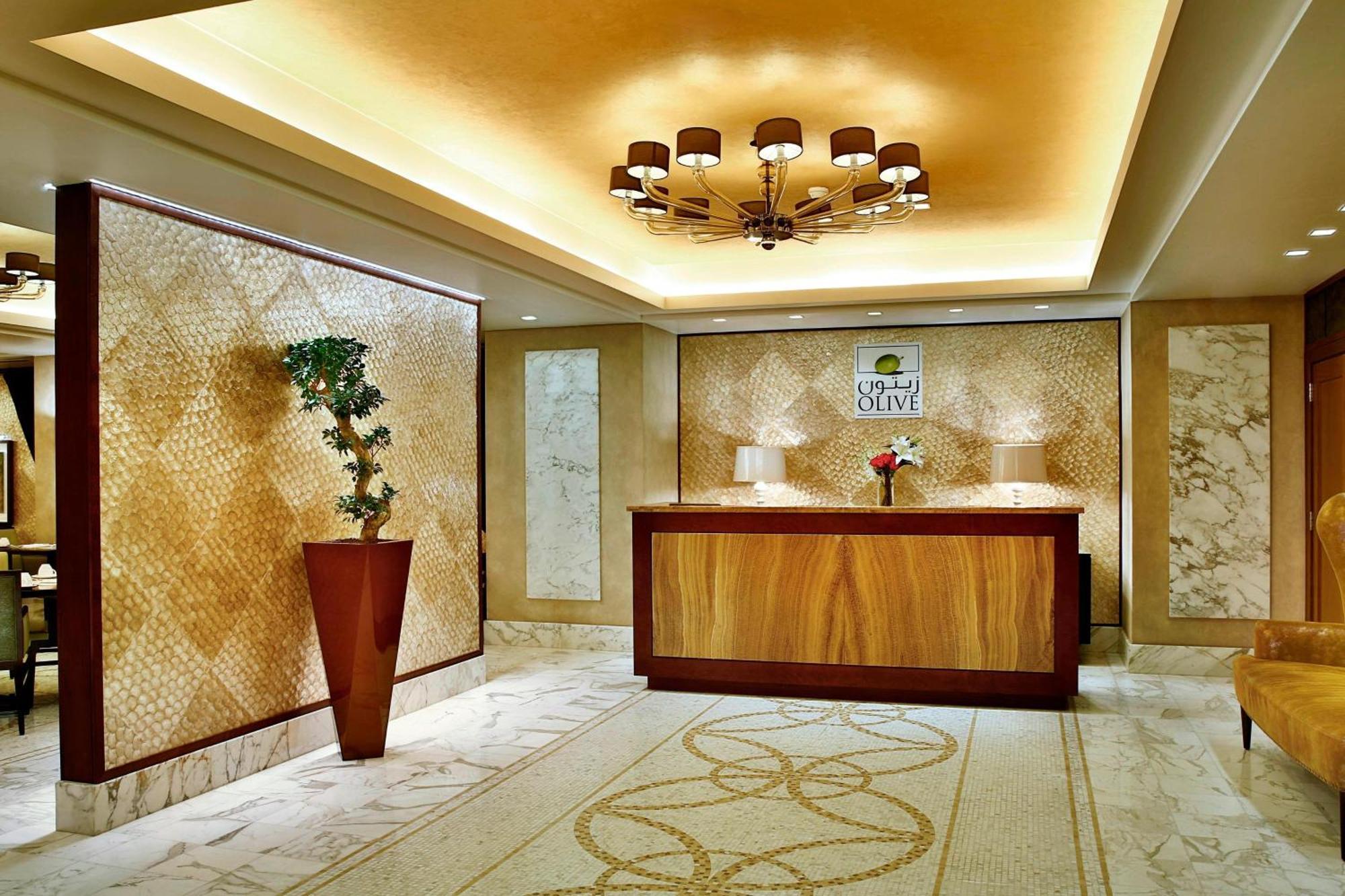 Jabal Omar Marriott Hotel Makkah Mecca Exterior photo The photo shows the interior of a hotel reception area. It features a wooden reception desk at the center, with a decorative wall behind it made of patterned materials. To the left, there is a tall potted plant adding a touch of greenery. The ceiling