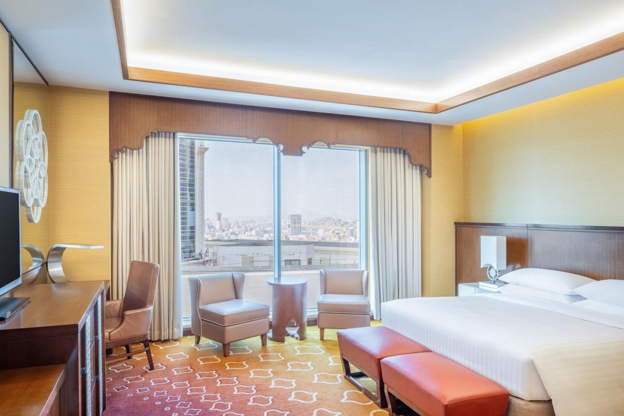 Jabal Omar Marriott Hotel Makkah Mecca Exterior photo The photo shows a modern hotel room featuring a comfortable bed with crisp white linens. There are two armchairs and a small table positioned by large windows that provide a view of the city. The room has warm, inviting colors with a patterned carpet