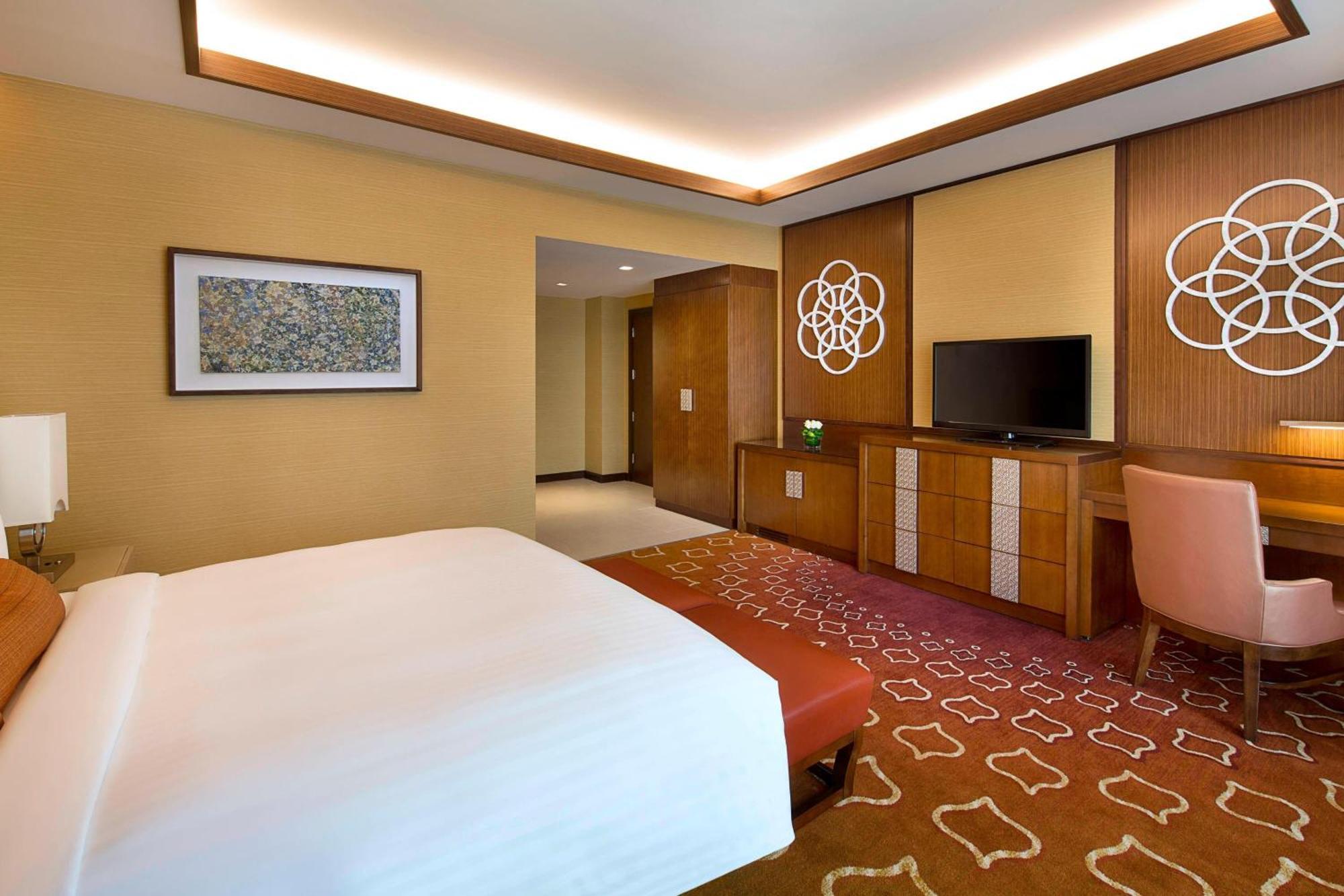 Jabal Omar Marriott Hotel Makkah Mecca Exterior photo The photo depicts a modern hotel room featuring a king-sized bed with a white comforter. The room has warm tones, with creamy walls and a patterned carpet in shades of orange. There is a wooden dresser with a flat-screen TV above it. Decorative wall 