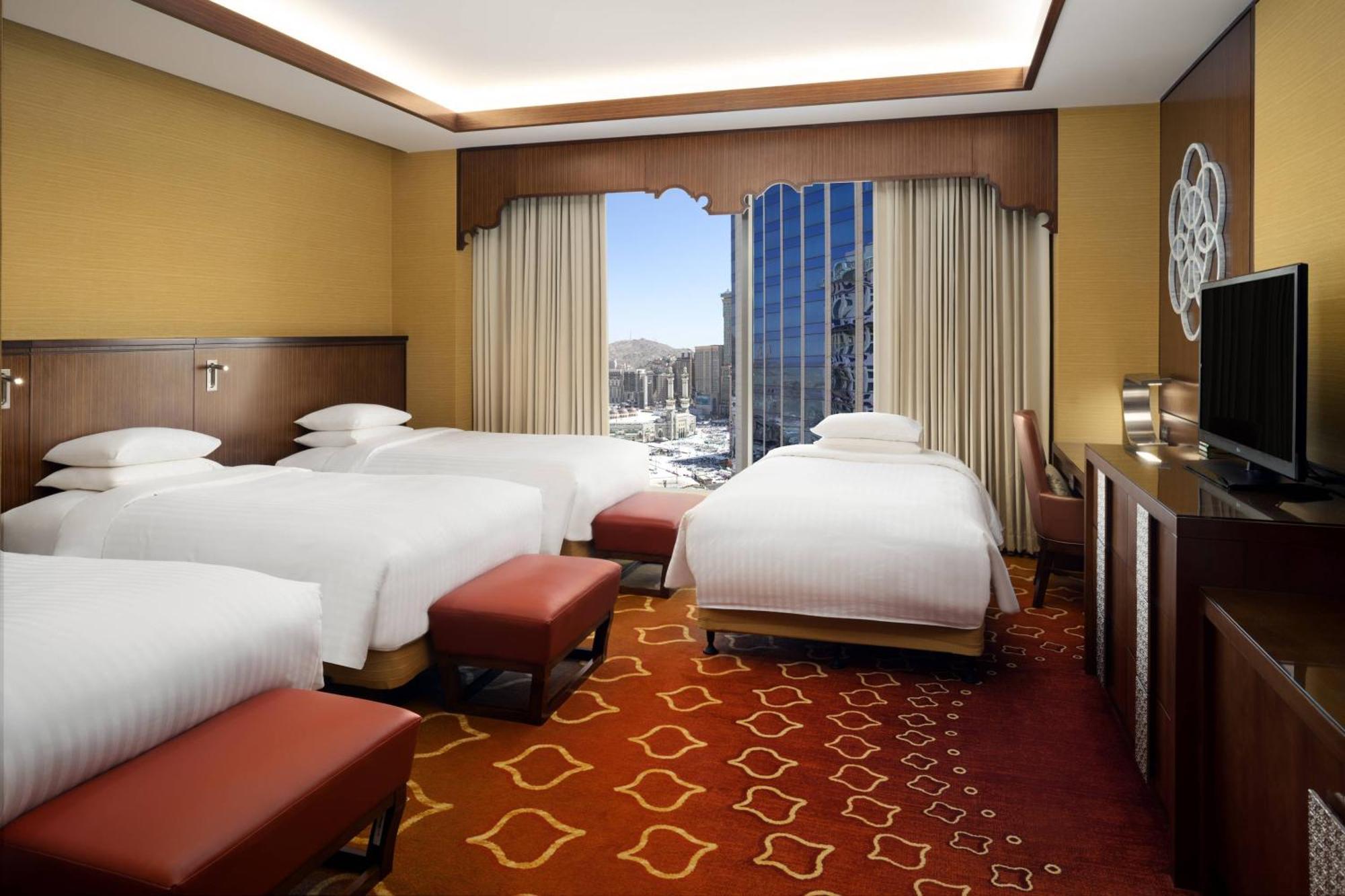 Jabal Omar Marriott Hotel Makkah Mecca Exterior photo The photo shows a hotel room featuring modern and stylish decor. There are three beds, each covered with white sheets, and reddish-brown benches positioned at the foot of the beds. The room has a large window with views of a cityscape outside, allowi